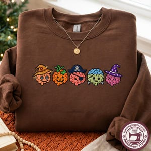 Puede incluir: Una sudadera marrón de cuello redondo con un bordado de cinco cerebros de dibujos animados disfrazados de Halloween, incluyendo un espantapájaros, una calabaza, un pirata, un monstruo de Frankenstein y una bruja, con un collar dorado encima.