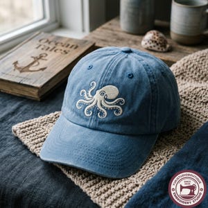 May include: A blue baseball cap featuring an embroidered cream-coloured octopus design on the front panel. The cap has a curved brim, a button on the top, and stitched eyelets for ventilation, showcasing a classic six-panel construction.