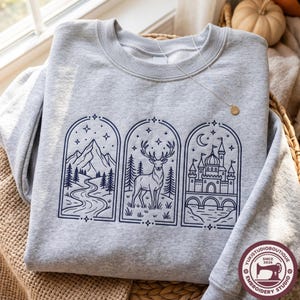 May include: A light gray crewneck sweatshirt featuring three vertical rectangular panels with dark blue line art. The panels depict a mountain landscape with a river, a deer standing in a forest, and a castle with a bridge under a crescent moon.