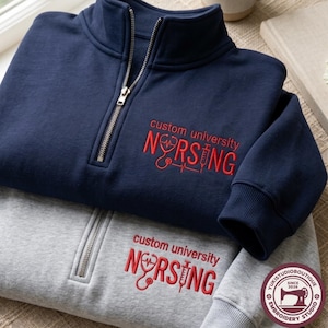 Puede incluir: Una sudadera azul marino y una gris claro con cremallera corta, ambas con bordado rojo que dice custom university NURSING junto a un gráfico de estetoscopio y jeringa. Las sudaderas tienen puños acanalados y un cierre de cremallera plateado.