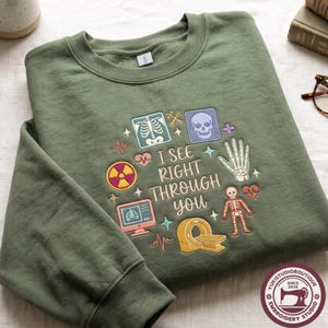 May include: An olive green crewneck sweatshirt featuring an embroidered design with the text "I SEE RIGHT THROUGH YOU" surrounded by medical icons including a skull, a hand skeleton, a full skeleton, an MRI machine, and a radiation symbol.