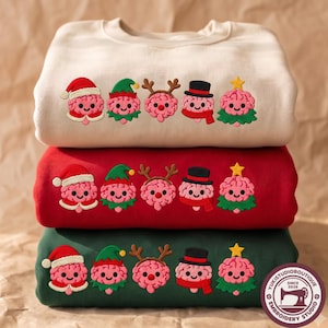 May include: A stack of three crewneck sweatshirts in cream, red, and dark green. Each features an embroidered row of five pink cartoon brains dressed as holiday characters: a Santa, an elf, a reindeer, a snowman, and a Christmas tree.