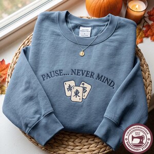 Könnte beinhalten: Ein staubblaues Sweatshirt mit Rundhalsausschnitt, bestickt mit dem Text PAUSE... NEVER MIND. und drei Mahjong-Steinen mit chinesischen Schriftzeichen. Eine goldfarbene Anhängerkette liegt am Ausschnitt und ein weißes Etikett zeigt COTTON ESSENTIALS.