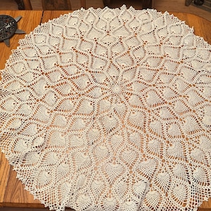 May include: A round, off-white crochet tablecloth featuring an intricate, repeating pineapple-inspired motif that radiates from the center outward to a scalloped edge. The delicate, openwork lace pattern is crafted with fine thread in a circular design.