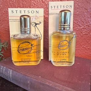 May include: Two bottles of Stetson cologne and spray are displayed. The bottles are clear with gold-colored liquid and silver caps. The packaging includes the Stetson logo and product information. The bottles are placed on a vintage book.
