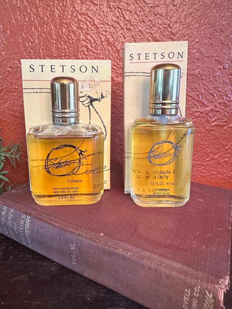 May include: Two bottles of Stetson cologne and spray are displayed. The bottles are clear with gold-colored liquid and silver caps. The packaging includes the Stetson logo and product information. The bottles are placed on a vintage book.
