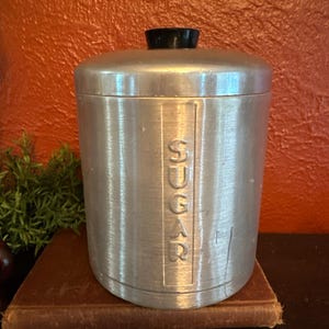 May include: A vintage metal sugar canister with a brushed finish. The word "SUGAR" is vertically embossed on the front. It has a black knob on the lid. The canister is cylindrical and sits on a brown surface.