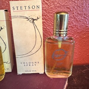 May include: Stetson cologne spray bottle and box. The box is tan with the Stetson logo and a drawing of a hand holding a rope. The bottle is clear with a gold cap and a blue logo. The box reads "COLOGNE SPRAY 1.5 FL OZ 44 mL".