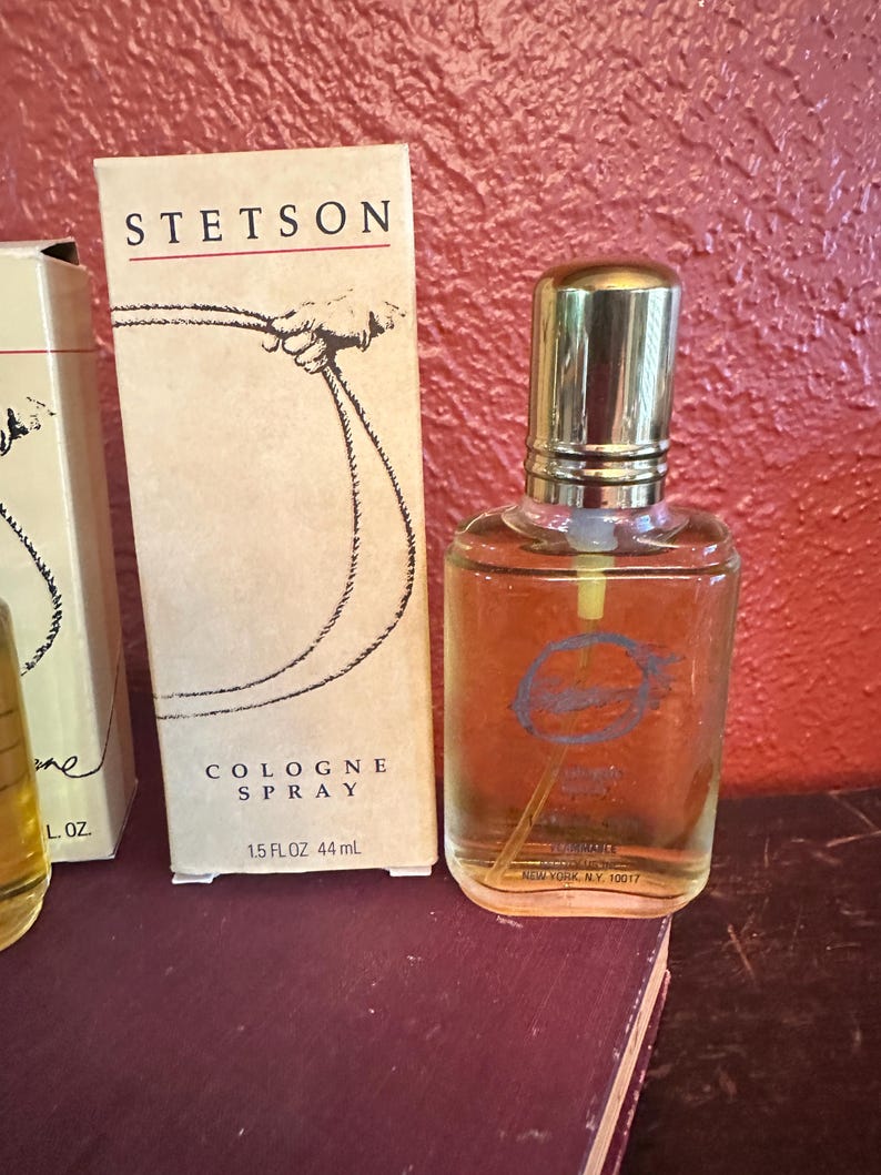 May include: Stetson cologne spray bottle and box. The box is tan with the Stetson logo and a drawing of a hand holding a rope. The bottle is clear with a gold cap and a blue logo. The box reads "COLOGNE SPRAY 1.5 FL OZ 44 mL".