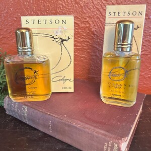 May include: Two bottles of Stetson cologne with gold caps and amber liquid, alongside their original boxes. The boxes feature the Stetson logo and a rope design. One bottle is 2.0 fl oz, the other is 1.5 fl oz. The bottles rest on a vintage book.