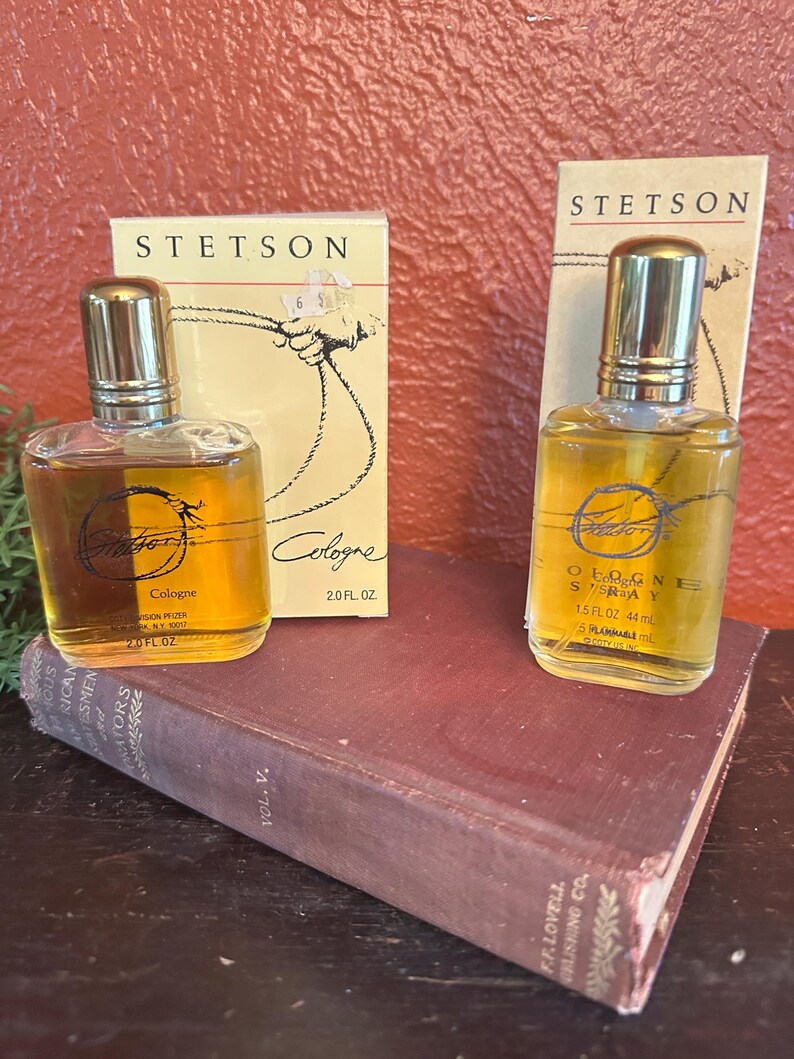 May include: Two bottles of Stetson cologne with gold caps and amber liquid, alongside their original boxes. The boxes feature the Stetson logo and a rope design. One bottle is 2.0 fl oz, the other is 1.5 fl oz. The bottles rest on a vintage book.