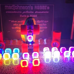 May include: A neon-lit display of custom roller skate wheels and a skateboard. The wheels and skateboard are illuminated with vibrant colors. A sign in the background reads "madjohnson's ROSEE's custom plate dreams".