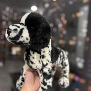 Op de afbeelding: Een pluche hondenspeelgoed met een zwart-wit gevlekt patroon. Het speelgoed heeft zwarte oren en een zwarte vlek over de ogen. De hond staat op alle vier de poten en lijkt van zacht materiaal te zijn gemaakt.