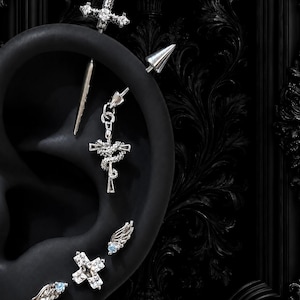 May include: A silver industrial barbell featuring a sword-shaped top and a dangling snake-wrapped cross charm. Below, a cross-shaped stud with clear crystals is flanked by two small wing-shaped studs with light blue stones on a black ear model.