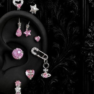 May include: A collection of silver-toned ear piercings featuring pink gemstones in various shapes including hearts, stars, and a circular stud. One piece is a safety pin design with a dangling orb charm. All items are displayed on a black ear model.