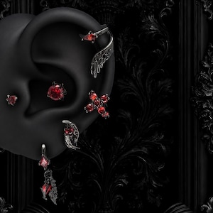 May include: A selection of dark silver earrings and ear cuffs, each featuring bright red gemstones. The collection includes a cross, wing-shaped designs, and dangling pieces, set against a black backdrop.
