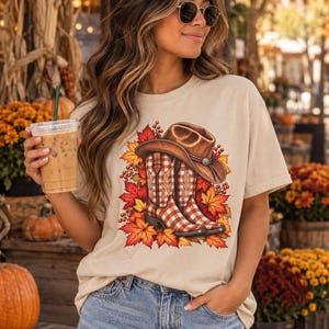 Könnte beinhalten: Ein beiges Kurzarm-T-Shirt mit einem Grafikdruck eines braunen Cowboyhuts, der auf einem Paar rot-weiß karierten Cowboystiefeln ruht, umgeben von einer Ansammlung leuchtend orangefarbener, gelber und roter herbstlicher Ahornblätter.