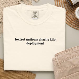 Op de afbeelding: Crèmekleurig T-shirt met de zwarte tekst "foxtrot uniform charlie kilo deployment". Het shirt is opgevouwen, met een beige broek ernaast. Het T-shirt heeft een Comfort Colors label. De achtergrond is neutraal.