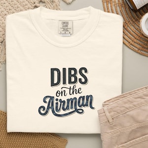 May include: Cream-colored t-shirt with the text "DIBS on the Airman" in a dark blue, vintage-style font. The shirt is folded, with a pair of tan pants visible. A woven beige mat and a small candle are in the background.