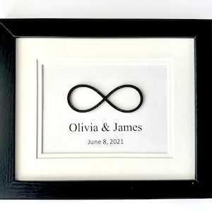 May include: Framed artwork featuring a black infinity symbol on a white background, with the names "Olivia & James" and the date "June 8, 2021" printed below. The artwork is set within a white mat and a black wooden frame.