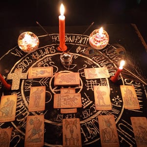 May include: A mystical scene featuring tarot cards arranged on a dark surface with a circular design. Lit candles and decorative glass orbs surround the cards. A glittery cross and peacock feather add to the ambiance.