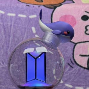May include: A light stick featuring a clear spherical top containing a blue illuminated BTS logo, topped with a small purple and white whale figure. The handle is black with a white geometric base and a small blue glowing logo on the front section.
