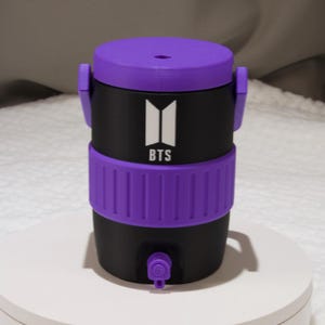 May include: A black beverage cooler with a purple lid, a purple textured middle band, and a purple spigot at the base. The front features a white logo with the text BTS. The cooler has two side handles and a cylindrical shape with a flat top.