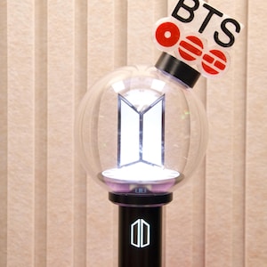 May include: A black and clear spherical light stick featuring a white BTS logo inside the globe. A black handle displays a white logo, and a white attachment at the top reads BTS with red circular accents. The device emits a bright white light.