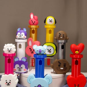May include: A collection of colorful plastic character-themed candy dispensers featuring various shapes and faces including hearts, clouds, and animals in shades of red, blue, yellow, purple, white, and brown, arranged on a circular white stand.