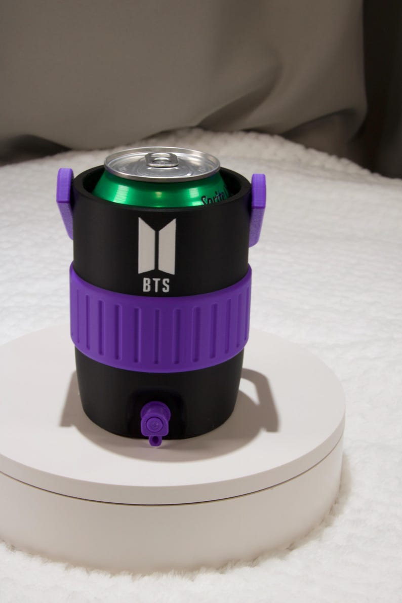 May include: A black cylindrical beverage can cooler featuring a purple ribbed band around the center and a small purple spout at the base. The front displays a white BTS logo with the text BTS underneath. A green soda can is inserted into the top.