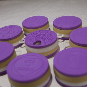 May include: A collection of round, cookie-shaped containers featuring purple lids with embossed designs, including the text OREO and BTS. Each container has a layered appearance with white and tan bands, and they are arranged on a white circular base.