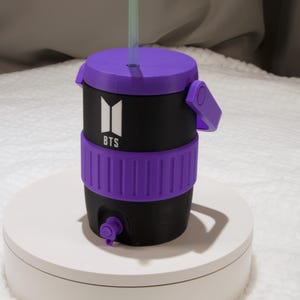 May include: A black and purple beverage cooler with a handle, a purple lid, and a straw. The front features a white BTS logo. The cooler has a purple textured band around the middle and a small purple spigot at the base for dispensing liquid.