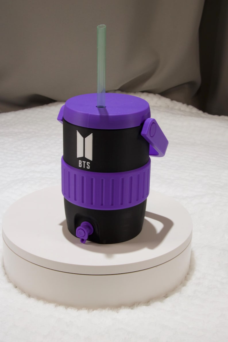 May include: A black and purple beverage cooler with a handle, a purple lid, and a straw. The front features a white BTS logo. The cooler has a purple textured band around the middle and a small purple spigot at the base for dispensing liquid.