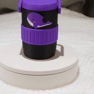 May include: A black cylindrical container with a purple lid and a matching purple silicone grip band. The container features a graphic of a purple whale with a white belly and a small pink cheek. The lid includes a purple handle and a small hole.