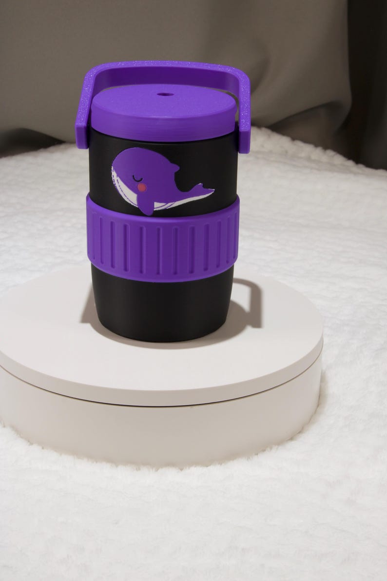 May include: A black cylindrical container with a purple lid and a matching purple silicone grip band. The container features a graphic of a purple whale with a white belly and a small pink cheek. The lid includes a purple handle and a small hole.