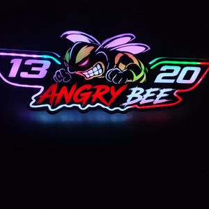 Peut inclure: Une enseigne LED lumineuse représentant une abeille de dessin animé stylisée avec une expression féroce, placée entre les chiffres 13 et 20. Le texte ANGRY BEE est affiché en lettres rouges grasses sous l'abeille, le tout sur fond sombre.