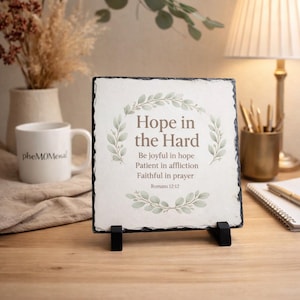 May include: Square slate plaque with the text "Hope in the Hard" and inspirational verses. The plaque is supported by a black stand and features a decorative wreath design. A white mug with the word "pheMOMenal" is visible.