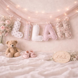 May include: A decorative banner spelling the name LILAH with individual fabric letters in various floral and solid pink and white patterns, accompanied by a light brown teddy bear, a pair of small pink baby shoes, and a wooden plaque with the name Lilah.