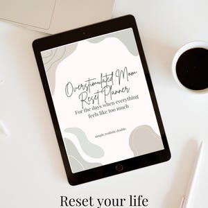 May include: A black tablet displaying a planner titled "Overstimulated Mom Reset Planner" with the tagline "For the days when everything feels like too much." The image includes a laptop, coffee cup, and pen.