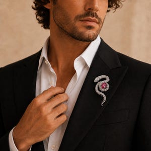 Puede incluir: Un broche de serpiente en tono plateado con una piedra preciosa ovalada de color rosa en el centro e incrustaciones de pequeñas piedras transparentes. El broche está prendido en la solapa de una americana negra sobre una camisa blanca.