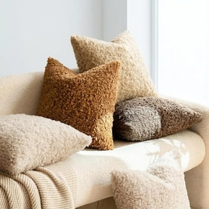 May include: A collection of textured throw pillows in various neutral tones, including cream, tan, brown, and gray. The pillows are arranged on a cream-colored sofa, creating a cozy and inviting aesthetic. The pillows have a soft, plush appearance.