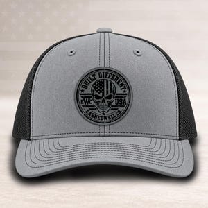 May include: A grey and black trucker hat featuring a circular patch on the front. The patch displays a skull with a flag pattern on its forehead, surrounded by the text BUILT DIFFERENT EWC USA. The hat has a solid grey front and black mesh back.