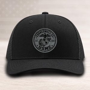 May include: A black baseball cap with a mesh back and a circular patch on the front. The patch features a dark gray eagle, globe, and anchor emblem with the text EARNEDWELL CO. EST. 2026 and USMC printed in a circular border design.