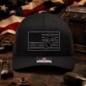 May include: A black trucker hat featuring a rectangular patch on the front. The patch displays a gray graphic of the American flag combined with the USMC Eagle, Globe, and Anchor emblem. A small rectangular sticker is centered on the brim.