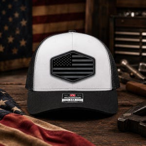 May include: A white and black trucker hat featuring a black hexagonal patch on the front with a monochromatic American flag design. The hat has a black brim and a mesh back, with a rectangular sticker on the brim reading Richardson 112.