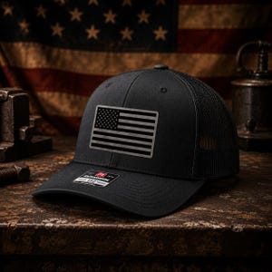 May include: A black trucker hat featuring a rectangular patch with a monochromatic American flag design on the front panel. The cap has a structured crown, a curved brim, and a mesh back. A small rectangular sticker is visible on the brim.