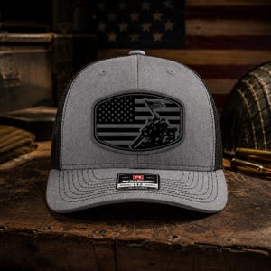 May include: A grey and black trucker hat featuring a rectangular patch on the front. The patch displays a black graphic of the American flag and the Iwo Jima flag raising. A small black and white sticker on the brim reads Richardson 112.