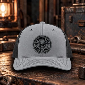 May include: A grey trucker hat with a black mesh back and a circular patch on the front. The patch features a skull graphic, the text PUNCH IN GRIND OUT, EWC, and EST 2026. The hat has a structured crown, a curved brim with stitching, and a button on top.