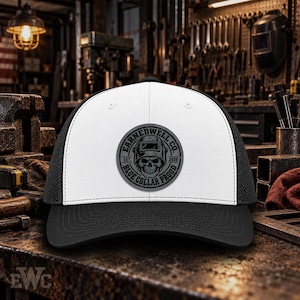 May include: A white and black trucker hat featuring a circular grey patch on the front. The patch displays a graphic of a skull wearing a welding helmet with the text EARNEDWELL CO. EST. 2026 and BLUE COLLAR PROUD printed around the border.