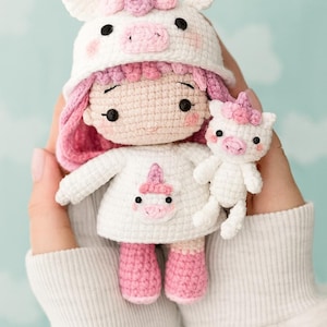 May include: A handmade crochet doll wearing a white dress and a unicorn hat with pink accents. The doll has pink boots and a matching small unicorn companion. The doll is held in front of a light blue background.